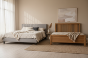 A soft grey upholstered platform bed and a solid oak wooden bed frame side by side in a bright warm-neutral bedroom.