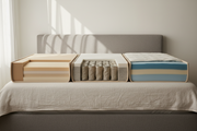 Three mattress cross-sections side by side — memory foam, pocket sprung and hybrid — on a neutral upholstered bed in soft daylight.