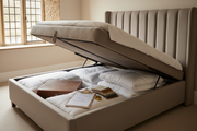 Taupe upholstered ottoman bed with gas-lift base raised open, showing lined storage compartment, with a checklist, tape measure and fabric swatches on the mattress
