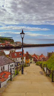 Stairway leading down to a coastal town with houses and a lamp post, under a cloudy sky.